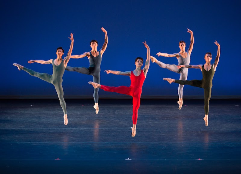 A male quintet, each in a primary color unitard jump off one leg. The other is pointed out the side. Their left arm is above their head.